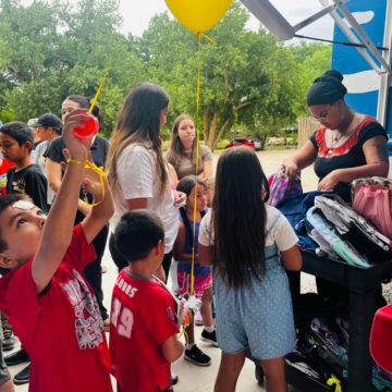 A crowd of young people, two holding balloons, line up as they receive backpacks.