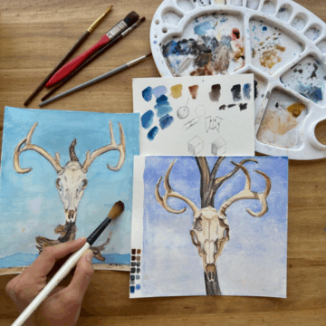 Two small paintings of a skull against a blue sky rest on a table. Above the paintings is a hand holding a paintbrush and above rests a palette.