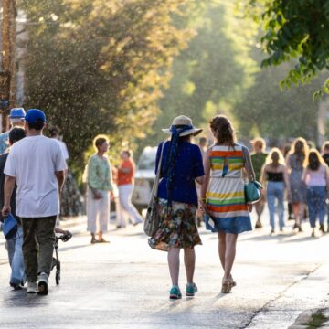A crowd of people walk down a street in the Summer sunlight. Green trees line the street.