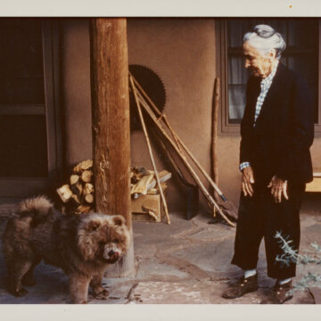 Photograph of Georgia O'Keeffe standing in her home courtyard with a small brown Chow Chow. O'Keeffe's grey hair is tied up and she wears a black and white suit.