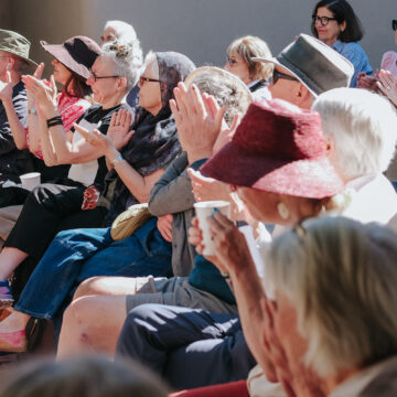 A group of people in an audience sit outdoors and clap. Several of them are wearing hats and sunglasses.