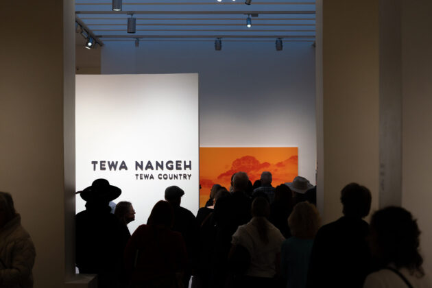 A group of people in front of an exhibition with the title 'Tewa Nangeh/Tewa Country' on a white wall. Behind is a large canvas of a billowing cloud in shades of orange.