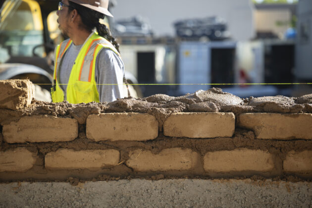 An adobe wall being built atop a concrete structure. Behind the wall stands a construction work in the background.