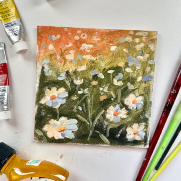 A small canvas with oil paintings of white daisies against a green and orange background. Surrounding the small canvas are paint tubes and several paintbrushes.