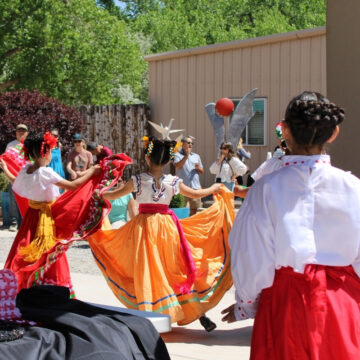 A group of young dancers with colorful large skirts perform in front of a crowd outside.