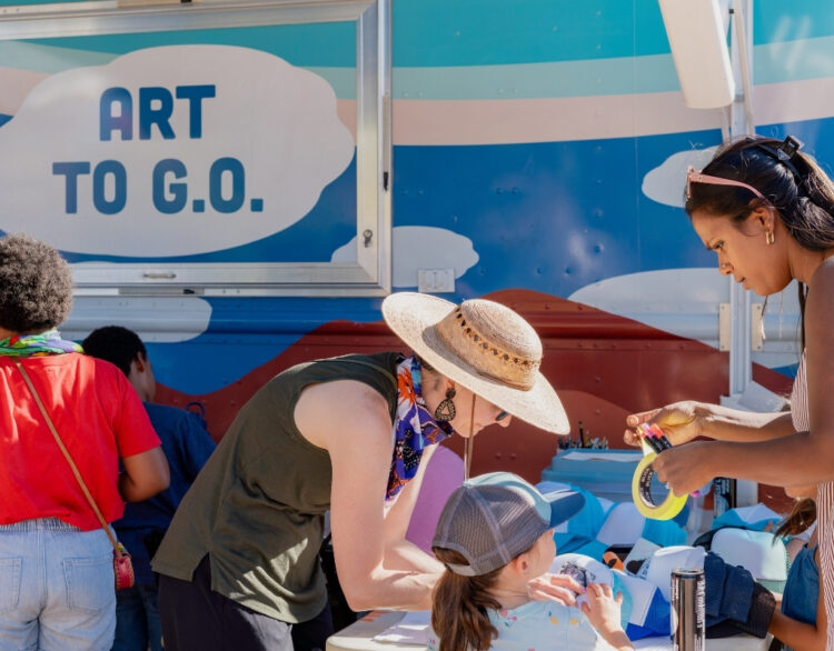 A group of adults and children stand over a table creating with their hands. Behind is a large truck with the label "Art to G.O"