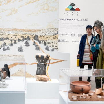 Photograph of people looking down onto a clay drum presented in an exhibition space. Behind is a large mural depicting a desert landscape.
