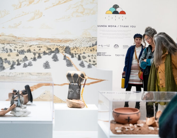 Photograph of people looking down onto a clay drum presented in an exhibition space. Behind is a large mural depicting a desert landscape.
