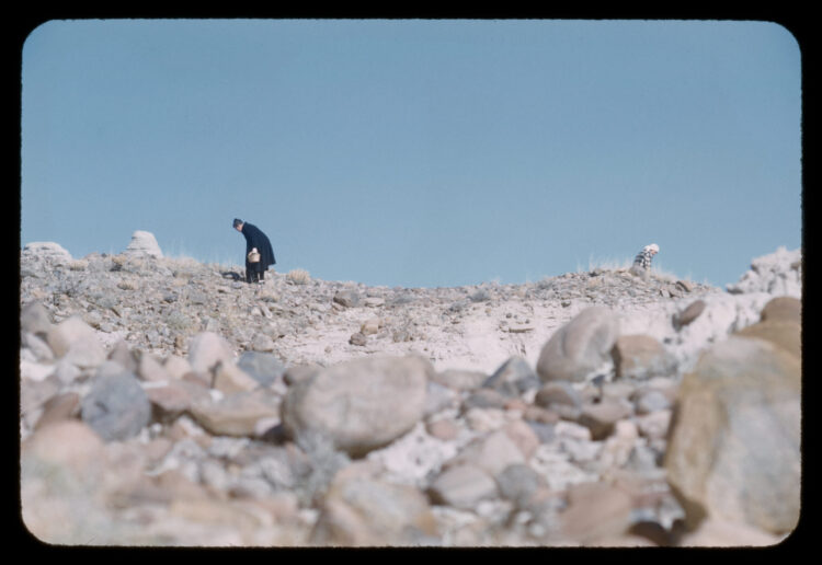 Photograph of two people very small and bent over looking at rocks against a blue sky.