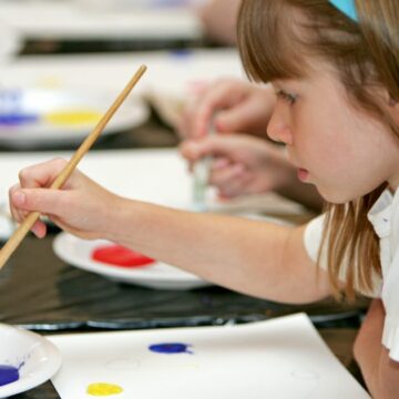 This photograph shows the side profile of a child holding a long paint brush. The tip of the paint brush is dipped into red ink. Her long hair is held back by a blue headband.