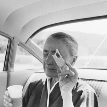 Portrait of Georgia O'Keeffe sitting in the backseat of a car holding a half full plastic cup in her right hand while holding piece of Swiss cheese up to her eye with her left hand; half-length view of O'Keeffe facing the front seat and with her right eye closed and her left eye looking towards the camera through the hole in the Swiss cheese ( like looking through the viewfinder of a camera or a pelvis opening in her paintings); her hair is pulled back from her face in a bun and she wears a black cotton wrap dress over a white cotton wrap dress.