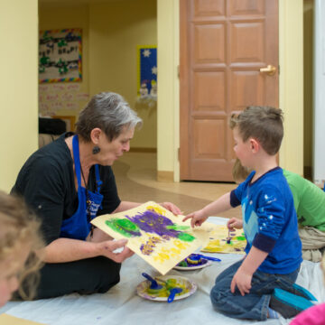 A toddler aged child in a blue shirt sits on his knees and pints to his green and purple artwork. A woman with short grey hair holds the painting and admires the child's work.