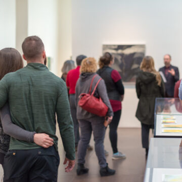 In the foreground we see the backs of a couple embracing in a side hug. The Georgia O'Keeffe Museum gallery is in the background. Other Museum visitors are in the background listening to a lecture.