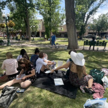 A group of students sit sketching on a green lawn with trees in the background.