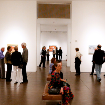 A photograph that depicts a group of 15 people milling about the Georgia O'Keeffe Museum. The foreground shows a woman in a floral dress sitting on a bench observing the artworks.