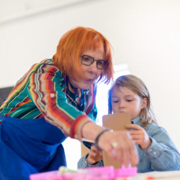 An adult in glasses and a child are bent over a piece of artwork on a table. The adult wears a blue apron.
